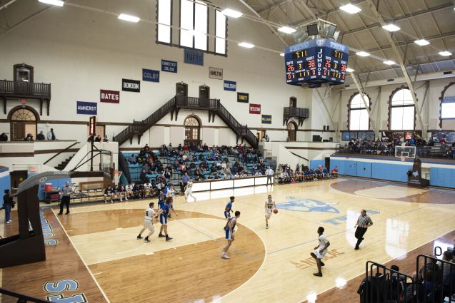 Medford和Somerville High Schools的男孩篮球队在阳光照射的塔夫茨品牌的健身房中互相玩耍，因为它充满了观众看起来。