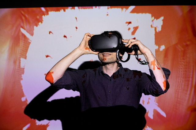 一个男人，戴着VR耳机覆盖着他的眼睛，反对橙色着色的屏幕。一所艺术博物馆教授和他的学生谈论VR的巨大潜力